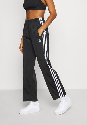 Femme portant un pantalon de survêtement Adidas noir avec des bandes blanches et le logo, des baskets blanches et un crop top noir, debout avec une main dans la poche.