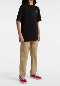 Černé tričko s krátkým rukávem a malým bílým logem na hrudníku, doplněné khaki kalhotami a červenými teniskami Vans.