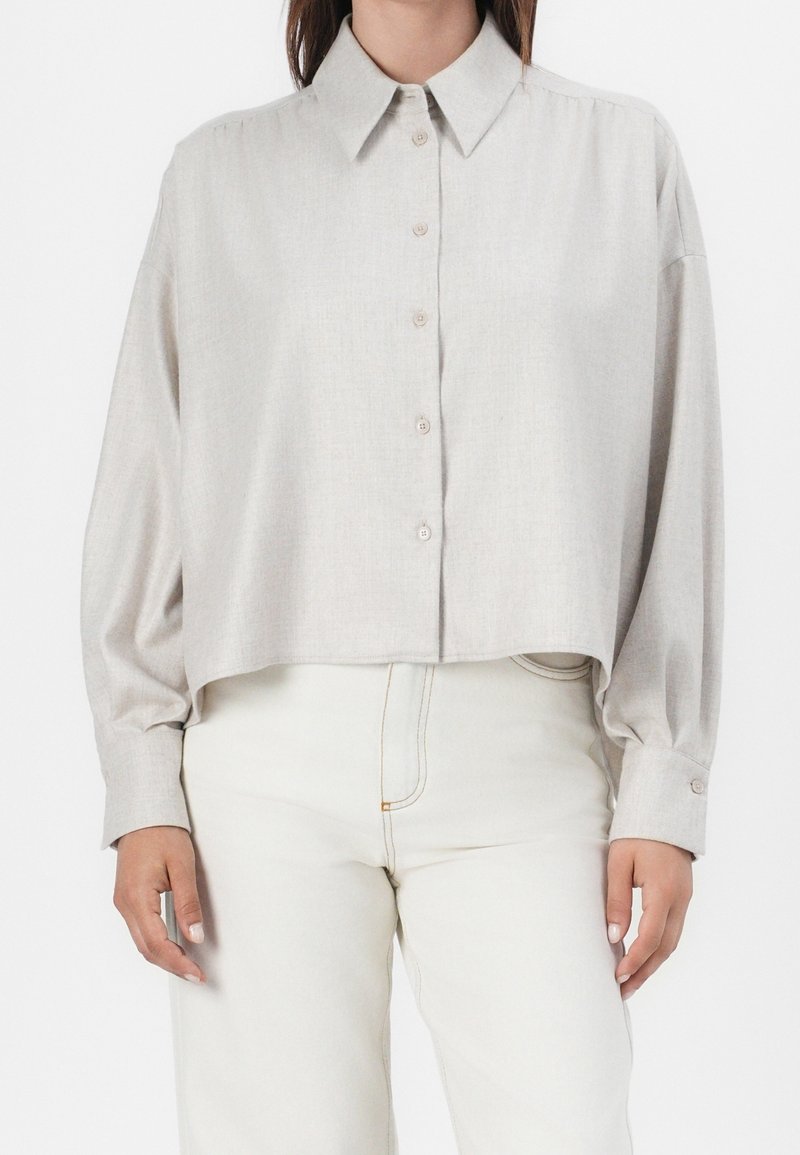 Femme portant une chemise beige à manches longues boutonnée avec un large col et un pantalon blanc taille haute, debout devant un fond blanc.
