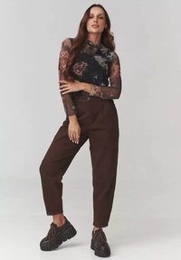 Kobieta stoi z jedną ręką skrzyżowaną na klatce piersiowej, ubrana w długi rękaw koronkowy top w kwiaty, brązowe spodnie z wysokim stanem oraz czarne buty na platformie.