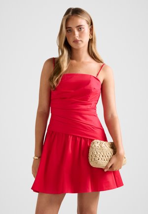 Joven mujer con un vestido rojo sin mangas, sosteniendo un bolso de mano beige tejido, llevando pendientes dorados y una pulsera, sobre un fondo claro y liso.