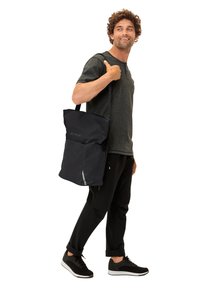 Schwarze Tragetasche mit strukturiertem Außenmaterial, ausgestattet mit einem oberen Verschluss, verstellbarem Schultergurt und reflektierenden Akzenten, getragen von einem männlichen Model.