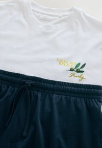 Fehér póló sárga „Valle del Belice” felirattal és zöld olívaág grafikával, sötétkék húzózsinóros nadrág felett.