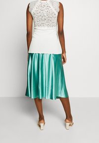 Kvinna i ärmlös vit spetsblus, tealfärgad satin midi-kjol och beige klacksandaler, stående mot en enfärgad ljus bakgrund.