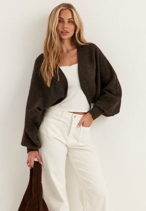 Blond kvinde iført en mørkebrun kort cardigan, hvid top og hvide bukser, der holder en matchende brun tekstureret taske.