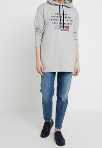 Sweat-shirt à capuche gris clair avec un texte graphique et un petit emblème rayé. Associé à un jean bleu délavé et des baskets noires.