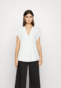 Weißes kurzärmliges Blusenshirt mit V-Ausschnitt, aus einem glatten Stoff; hat eine lockere Passform und dezente Seitenschlitze am Saum.