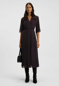 Nainen tumma purppuranvärinen, plissee-mallinen midimekko, jossa kyynärpään mittaiset hihat, mustat saappaat ja musta käsilaukku, yksiväristä taustaa vasten.