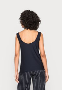 Kobieta o kręconych włosach, ubrana w bezrękawnik w kolorze czarnym i czarne spodnie w prążki, stojąca tyłem na jednolitym tle.