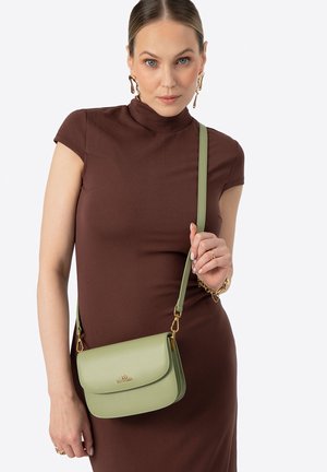 Kobieta w brązowej sukience i złotych kolczykach, trzymająca jasnozieloną skórzaną torebkę na ramię ze złotymi elementami.