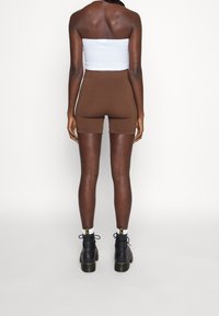 Personne debout, portant un haut blanc sans bretelles, un short marron ajusté, des chaussettes blanches et des bottes noires à lacets, face à un fond blanc uni.
