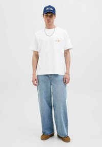 Witte katoenen t-shirt met een kleine oranje zonafbeelding, gecombineerd met lichtblauwe wijde denim jeans en bruine suède instapschoenen. Blauwe pet.