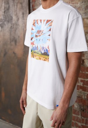 Henkilö, jolla on yllään valkoinen lyhyttakkinen paita, jossa on värikäs luontokohtaus ja "Nike Air" -logo, seisoo tiili- ja teollisuusseinää vasten.