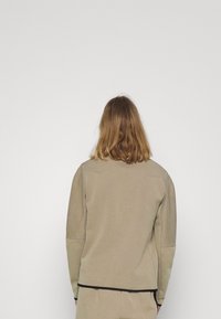 Person mit schulterlangem, hellbraunem Haar, der ein beigefarbenes Langarmoberteil und passende Hosen trägt, mit dem Rücken zu einer schlichten weißen Wand.