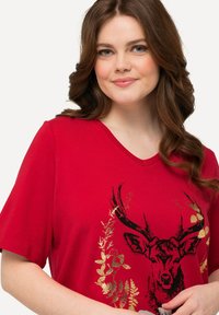 Camiseta de algodón roja con mangas cortas, escote en V, que presenta un gráfico de ciervo en negro y dorado con acentos florales. Textura suave y corte relajado.