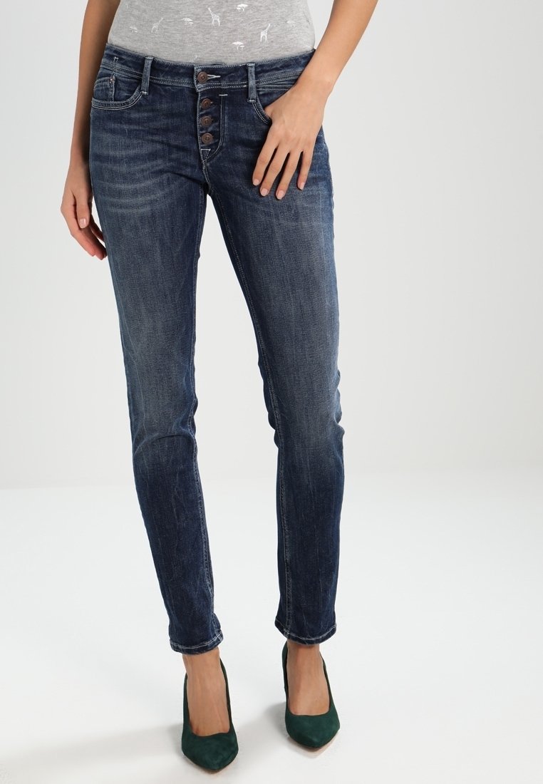 Jeans ajustados azules de denim con cinco bolsillos, cintura alta y cierre de botones. Llevados con tacones verde oscuro.