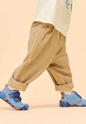 Bambino che cammina, indossando pantaloni beige risvoltati e scarpe blu con strappo, con dettagli arancioni e blu navy, su uno sfondo semplice.