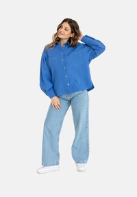 Niebieska, oversize koszula z guzikami, posiadająca dwie kieszenie z przodu, zestawiona z jasnoniebieskimi szerokimi spodniami i białymi trampkami. Gładka tekstura, swobodny fason.