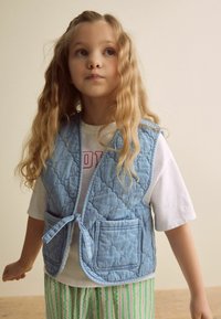 Helles blaues, gestepptes Weste mit Bindeschnürung und zwei Fronttaschen, getragen über einem weißen T-Shirt. Das Model hat lange, wellige Haare.