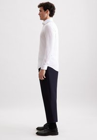 Uomo in profilo che indossa una camicia bianca a maniche lunghe, pantaloni blu scuro, calze nere e scarpe eleganti nere, su uno sfondo semplice.