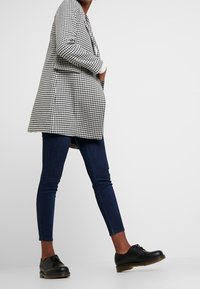 Cappotto lungo in bianco e nero a scacchi, abbinato a jeans skinny scuri e scarpe stringate nere, creando una silhouette strutturata.