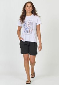 Balta t-kreklā ar krāsainu abstraktu apdruku un vārdu "BOHEMIAN", apvienots ar melniem šortiem un melnām flip-flops. Modelis stāv un staigā.