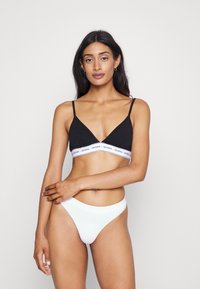 Black cotton bralette with thin straps and a white branded band; paired with white ribbed bikini bottoms. Simple, functional design.