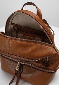 Brązowy skórzany plecak o teksturowanej powierzchni, ze złotymi suwakami i wewnętrzną kieszenią. Wewnątrz widoczne logo marki Michael Kors.