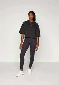 Γυναίκα που στέκεται φορώντας ένα μαύρο oversized T-shirt, μαύρες κολάν με λογότυπα και λευκά αθλητικά παπούτσια, μπροστά από έναν απλό φωτεινό φόντο.