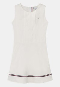 Ärmlös vit A-linjeklänning med plisserad framdel, liten broderad logotyp på bröstet och röda, vita och marinblå ränder nära fållen.