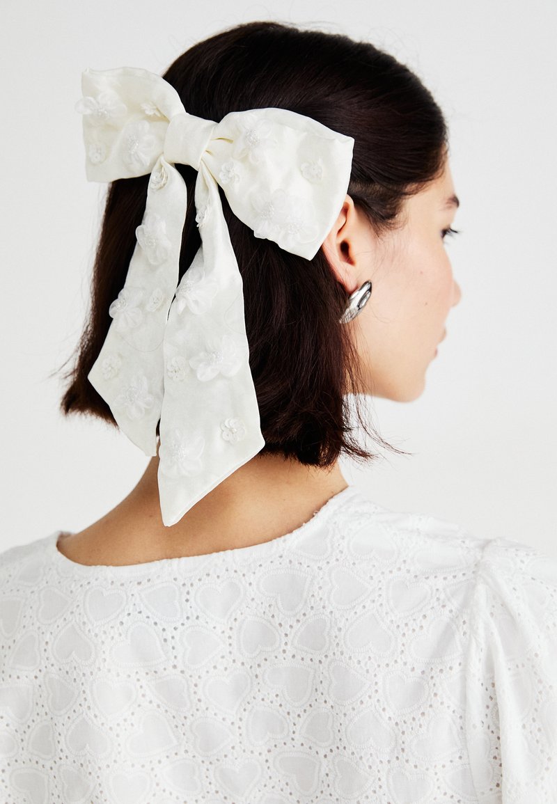 Fehér hajpánt, textúrált anyagból készült, virágmintás hímzéssel és lógható szalagokkal, laza hajban viselve, fehér szívmintás toppal párosítva.