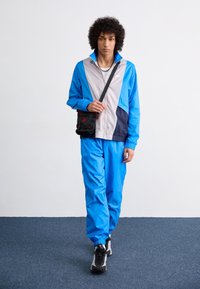 Modrá a šedá větrovka s elastickými manžetami, doplněná o odpovídající modré kalhoty a černé tenisky. Malá černá taška s červenými akcenty.