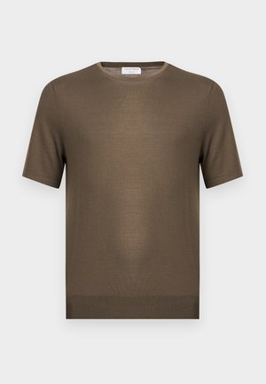 Νέος άνδρας με κατσαρά μαλλιά, φορώντας ένα ελιά κοντομάνικο μπλουζάκι, φαρδιά ξεβαμμένα μπλε τζιν και λευκά αθλητικά παπούτσια, στέκεται με το ένα χέρι στην τσέπη.