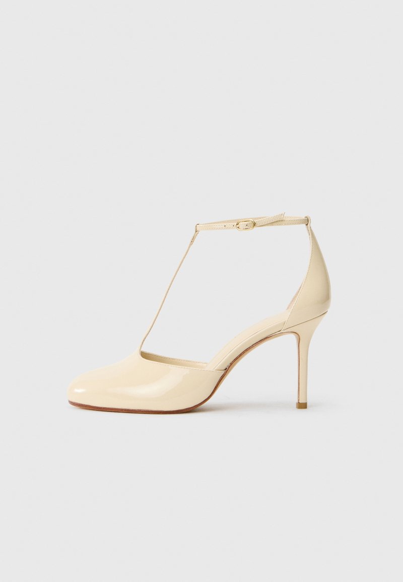 Pompe à talons hauts en cuir verni beige, avec une brides en T et une boucle à la cheville, un bout en amande et un talon aiguille. Fini lisse et brillant.