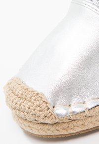 Espadrille métallique argenté avec une semelle en jute tressé. Présente un embout rond et une couture visible le long du bord de la chaussure.