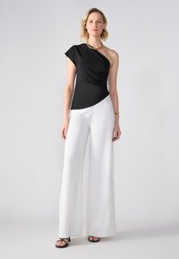 Top nero a spalla unica con design drappeggiato abbinato a pantaloni bianchi a gamba larga, completato da sandali neri con tacco e una collana d'oro.