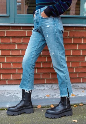 Persoon draagt lichtblauwe versleten cropped jeans met rafelige zomen en zwarte chunky plateau laarzen, staand tegen een bakstenen muur.