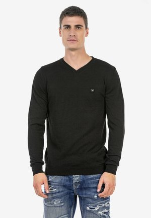Schwarzer V-Ausschnitt-Pullover, lange Ärmel, aus einem glatten Stoff. Kleines Logo-Patch auf der Brust, kombiniert mit verwaschenen blauen Jeans.