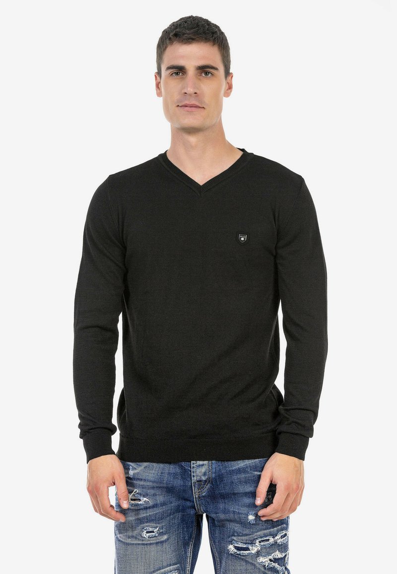 Schwarzer V-Ausschnitt-Pullover, lange Ärmel, aus einem glatten Stoff. Kleines Logo-Patch auf der Brust, kombiniert mit verwaschenen blauen Jeans.