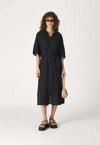 Black button-up dress with a waist tie, short sleeves, and a knee-length hem. Accessories include a small tan bag and black platform sandals.