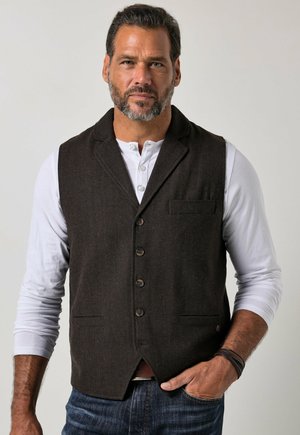 Man with beard wearing dark brown buttoned vest over white long-sleeve shirt and blue jeans standing with one hand in pocket.