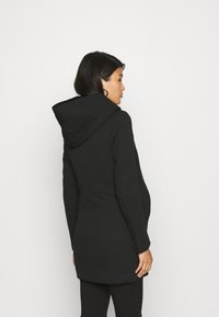 Manteau à capuche noir, mi-long, en tissu texturé. Présente un design ajusté avec des manches longues et des poches latérales discrètes.