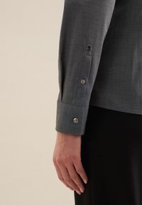 Manica di camicia grigia, strutturata, con polsino abbottonato. Presenta un motivo sottile e un dettaglio di fiore ricamato vicino al polsino.