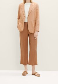 Hellbrauner Leinen-Blazer mit Ein-Knopf-Verschluss, kombiniert mit passenden weit geschnittenen Hosen und beigen Riemchensandalen. Glatte Textur insgesamt.