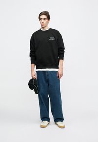 Črna majica s kapuco z logotipom, pod njo bela ovratna plast, ohlapne modre jeans hlače in bež superge. Drži črno kape z belimi detajli.