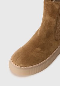 Bottine en suede marron avec des coutures décoratives et une épaisse semelle en caoutchouc beige sur fond blanc.