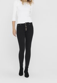 Vaqueros ajustados negros de talle alto, con herrajes de botones dorados, bolsillos delanteros y una textura de denim suave. Combinados con una blusa blanca.
