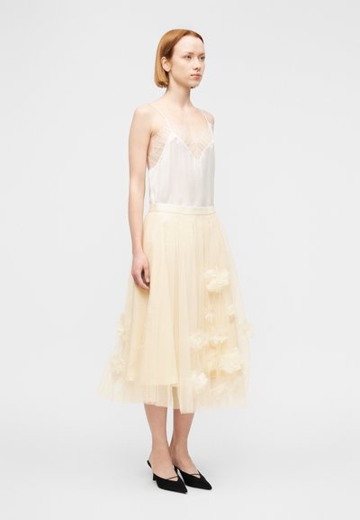 Ελεφαντόδοντο μεταξωτό καμιζόλ με δαντελένιο τελείωμα, συνδυασμένο με κίτρινη διαφανή φούστα από τούλι, πλισέ μήκους μέχρι το γόνατο, διακοσμημένη με ανάγλυφες εφαρμογές με λουλούδια. Μαύρα παπούτσια.
