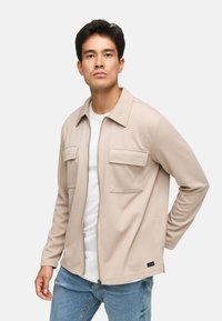 Beige zip-jakke med krave, to brystlommer og struktureret stof, båret over en hvid skjorte og parret med lyseblå jeans.