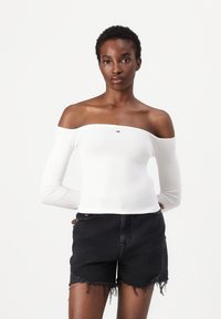 Bela, off-the-shoulder bluzo z dolgimi rokavi iz rebraste tkanine z manjšim logotipom na prsih, v kombinaciji z črnimi raztrganimi denim kratkimi hlačami.